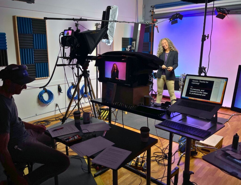 In a well-equipped studio, an individual sits monitoring equipment while another stands smiling before the camera, with colorful backdrops and professional lighting set up around them.
