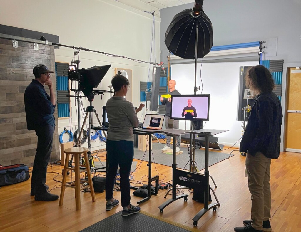 A behind-the-scenes photo of a video shoot at Studio V, showing three people working with professional video equipment, including a camera, lighting, and a monitor displaying the subject being filmed.