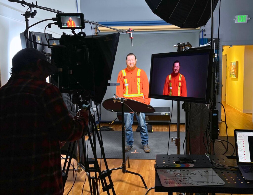 Image of a video production setup at Studio V featuring a cameraman focusing on a smiling subject in a high-visibility orange vest, with the camera's viewfinder and a large monitor showing the live shot. 