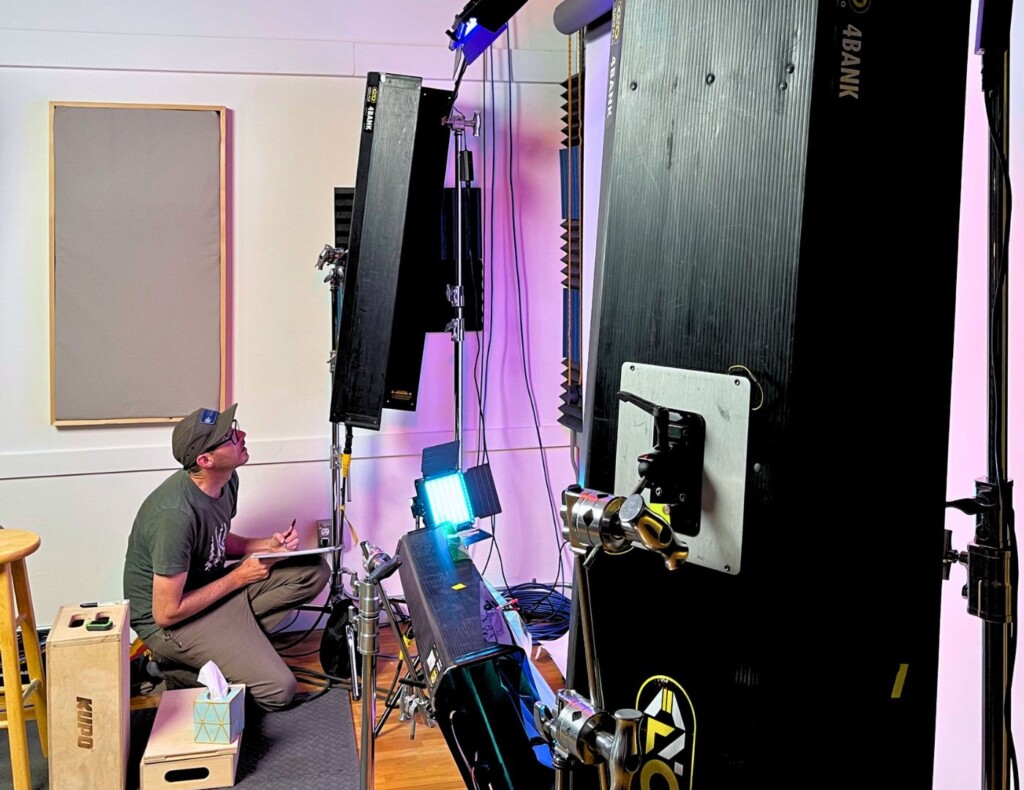 A man in a cap and glasses is adjusting a large vertical lighting panel in a studio setting, with various lighting equipment and a camera in the foreground.